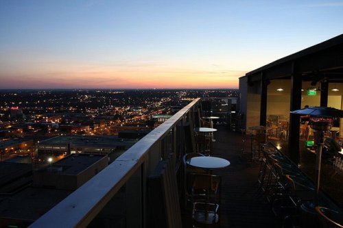Rooftop view at sunset, showcasing a city skyline with lights twinkling below and a few tables on the deck.
