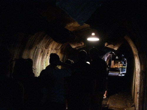 A dimly lit tunnel with people walking towards the light at the end, revealing machinery in the background.