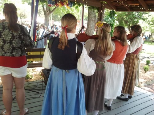A group of musicians in traditional attire perform on a stage under a gazebo, surrounded by trees and an audience.