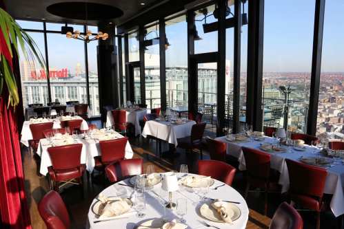 Elegant restaurant with floor-to-ceiling windows, featuring tables set for dining and a city skyline view.