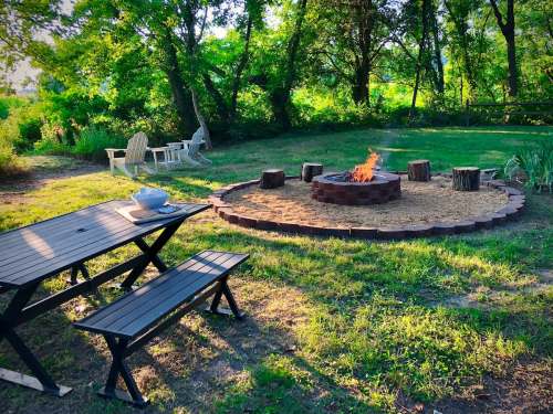 A cozy outdoor fire pit surrounded by logs, a picnic table, and chairs, set in a lush green garden.