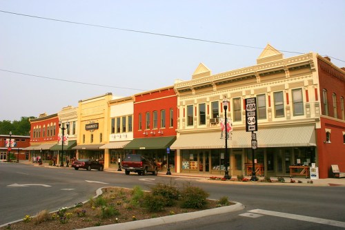 Historic storefronts line a street, featuring colorful facades and awnings, with parked cars and greenery in the foreground.