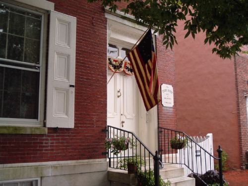 A brick building entrance with a white door, American flag, and decorative plants on the steps.