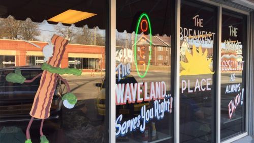 Colorful window display of a diner featuring a cartoon bacon character and signs promoting breakfast and the diner’s name.