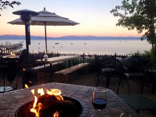 A serene lakeside view at sunset, featuring a fire pit, wine glass, and boats on the water.