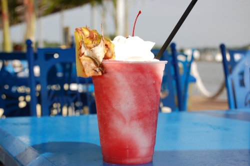 A vibrant red cocktail topped with whipped cream and a cherry, served in a clear cup on a blue table by the water.