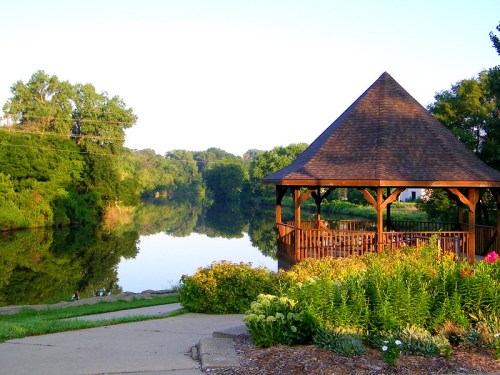 A wooden gazebo by a calm lake, surrounded by lush greenery and colorful flowers under a clear blue sky.