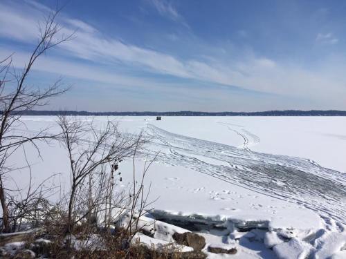 A snowy landscape with a frozen lake, featuring tire tracks and a small structure in the distance under a clear blue sky.
