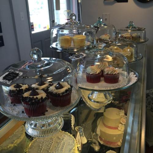 A display of assorted cupcakes under glass domes in a bakery, with a cake visible in the foreground.