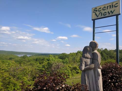 A stone statue of a couple embraces, overlooking a lush green landscape and a river under a blue sky.