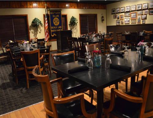 A cozy restaurant interior with dark wooden tables, chairs, and decorative plants, featuring a Rotary Club banner on the wall.