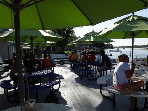 A lively outdoor seating area with green umbrellas, people chatting, and a view of the water in the background.