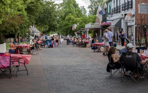 A lively outdoor street scene with people dining at tables, surrounded by shops and greenery on a sunny day.
