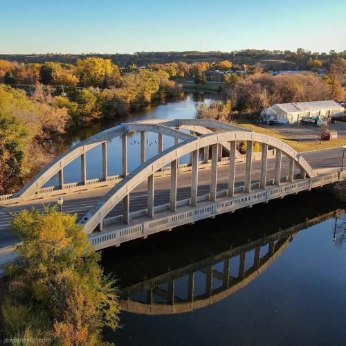 Aerial view of a curved bridge over a calm river, surrounded by autumn trees and a clear blue sky.
