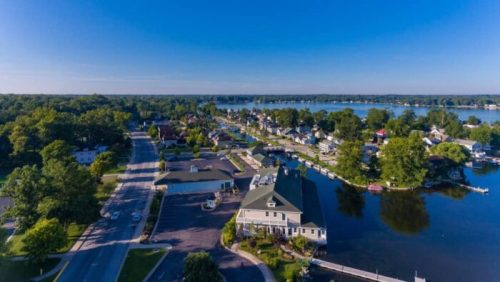 Aerial view of a serene lakeside community with homes, trees, and a calm waterway under a clear blue sky.