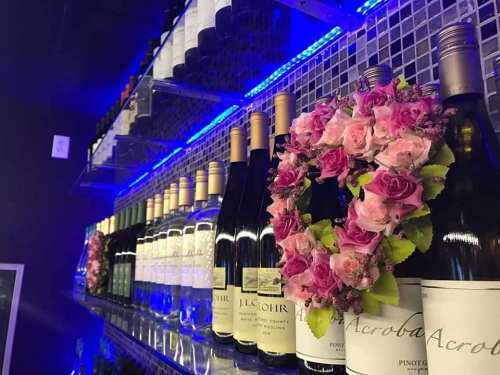 A shelf displaying various wine bottles, adorned with a floral arrangement of pink roses. Blue lighting enhances the scene.
