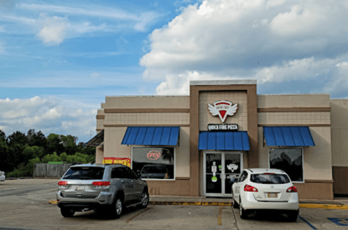 A pizza restaurant with blue awnings and a sign, surrounded by parked cars under a partly cloudy sky.
