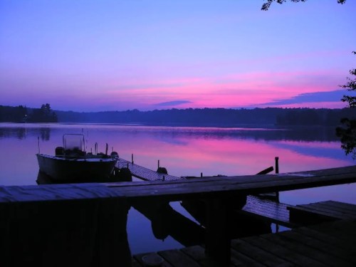 A serene lake at sunset, with vibrant pink and purple hues reflecting on the water and a dock in the foreground.