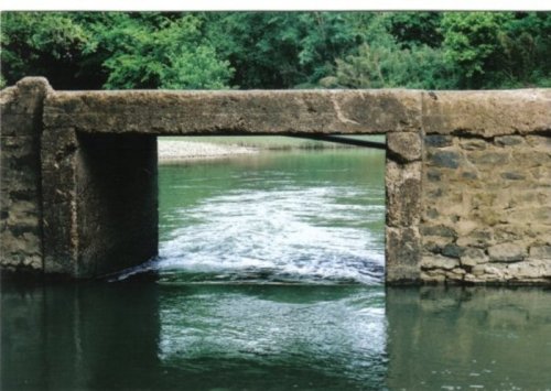 A stone bridge over a calm river, surrounded by lush greenery and trees. Water flows gently beneath the arch.