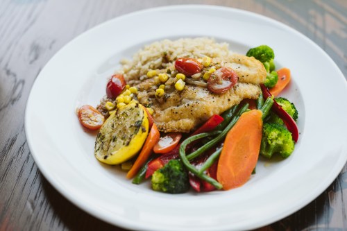 A plate of grilled fish with vegetables, quinoa, and lemon, garnished with cherry tomatoes and corn.