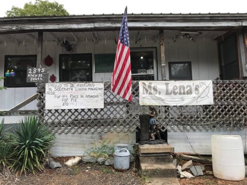 A rustic storefront with an American flag, signs for Ms. Lena's, and a welcoming atmosphere in a rural setting.