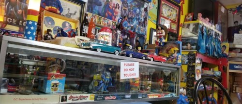 A colorful display of superhero memorabilia, including toys, posters, and collectibles, with a "Please do not touch" sign.