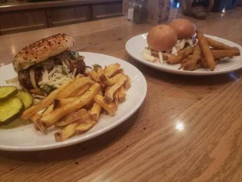 A plate with a burger topped with greens and pickles, alongside fries; another plate with sliders and fries in the background.