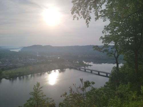A serene view of a river reflecting sunlight, surrounded by hills and trees, with a bridge in the distance.