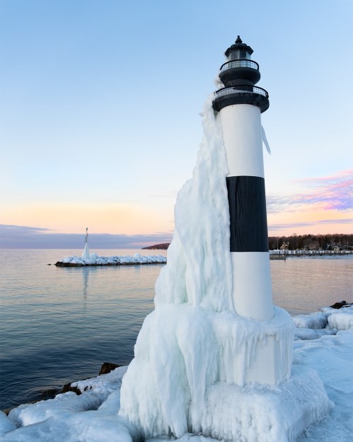 A frozen lighthouse stands by the water, surrounded by ice, with a colorful sky at sunset in the background.