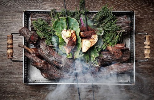 A tray with smoked food, including herbs and vegetables, surrounded by wooden elements and wisps of smoke.