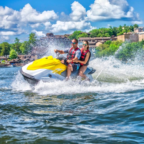 A man and woman ride a yellow jet ski on a lake, splashing water under a blue sky with fluffy clouds.