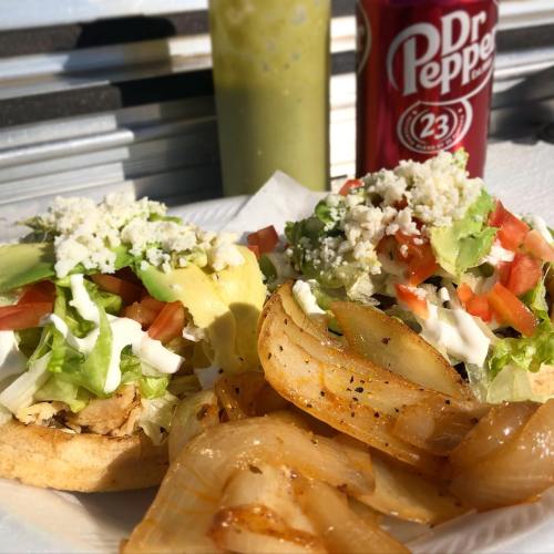 Two loaded tacos with chicken, lettuce, tomatoes, avocado, and cheese, served with grilled onions and a soda.