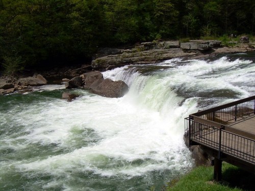 A waterfall cascades over rocks into a river, surrounded by lush greenery and a wooden viewing platform nearby.