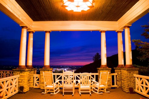 A scenic porch with white rocking chairs, overlooking a city skyline at dusk, illuminated by a warm light fixture.