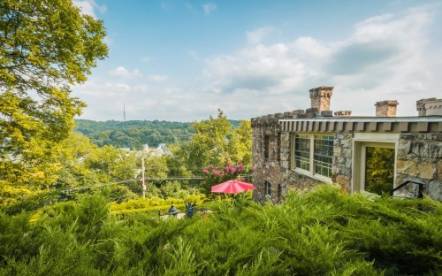 A scenic view of a stone house surrounded by greenery, with a red umbrella and hills in the background under a blue sky.