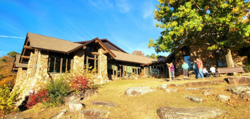 A rustic stone building surrounded by trees, with people gathered outside on a sunny day.