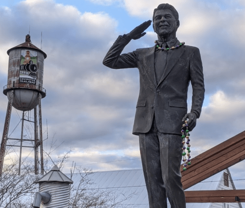 A statue of a smiling man in a suit saluting, with a water tower and cloudy sky in the background.