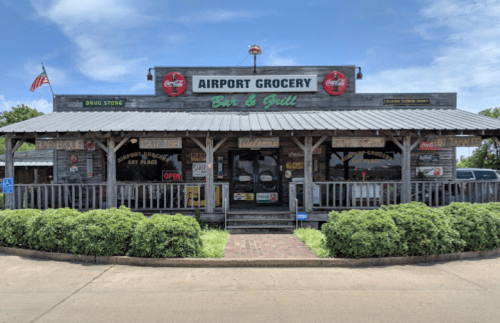 A rustic bar and grill named "Airport Grocery" with a wooden porch and various signs, set against a blue sky.