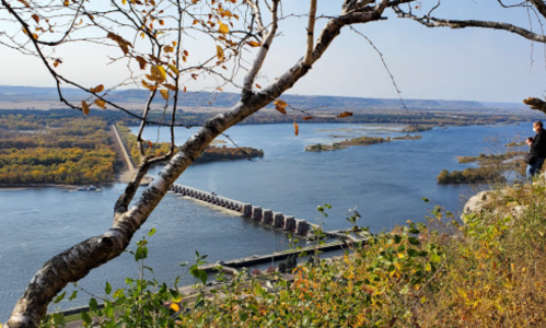 A scenic view of a river with a dam, surrounded by autumn foliage and a person standing on a rocky ledge.