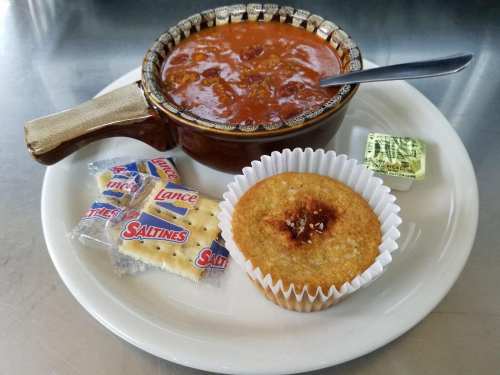 A bowl of chili with a spoon, served with crackers, a muffin, and a butter packet on a white plate.
