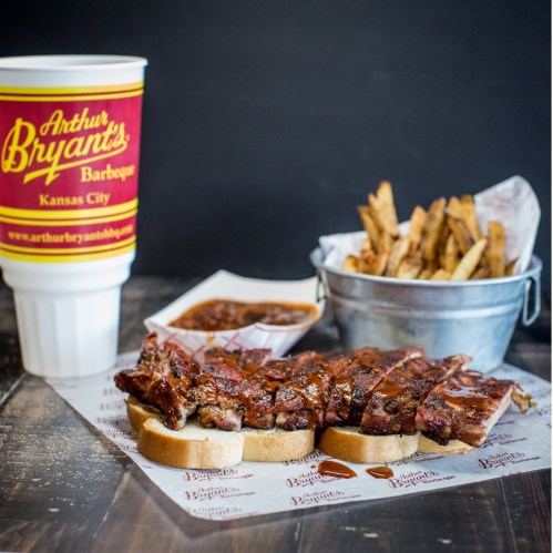 A plate of barbecue ribs on bread, a cup of drink, and a bucket of fries on a wooden table.