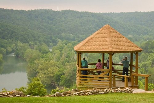 A wooden gazebo overlooks a river and lush green hills, with four people enjoying the scenic view.