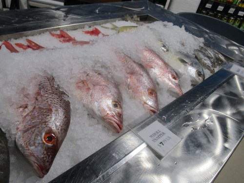 A display of various whole fish on ice at a seafood market, labeled with prices.
