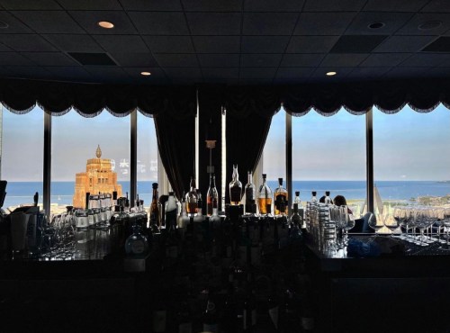 A bar with various bottles in front of large windows showcasing a coastal view and a distant building.
