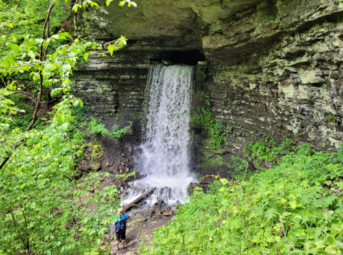 A person stands near a waterfall cascading over rocky cliffs, surrounded by lush green foliage.