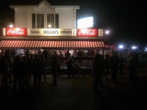 A busy ice cream shop at night, with a striped awning and a crowd enjoying sodas and sundaes.