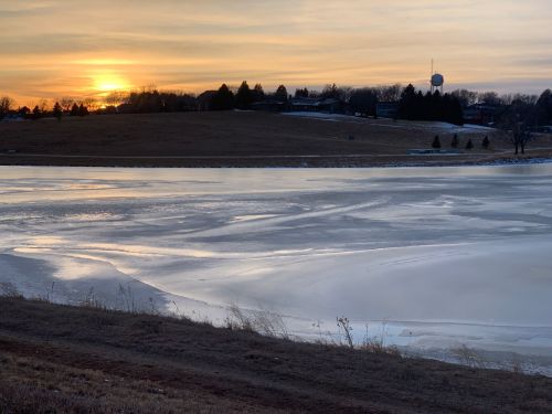 Sunset over a frozen lake, with a grassy shore and distant buildings silhouetted against the colorful sky.