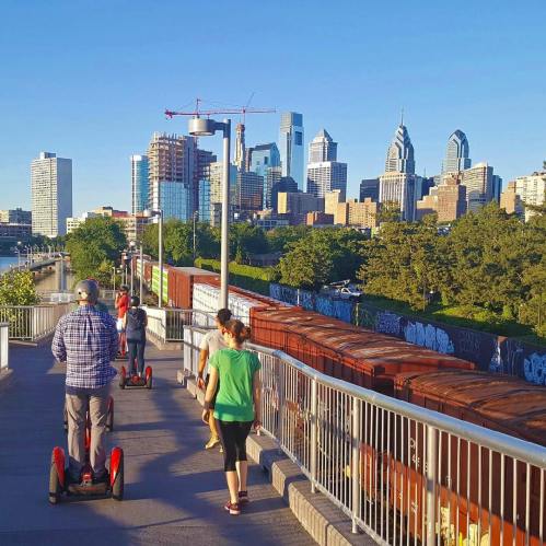 People riding segways along a waterfront path with a city skyline in the background on a sunny day.