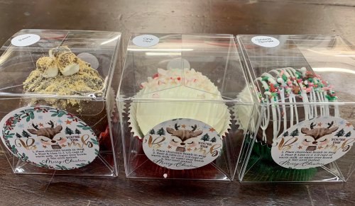 Three festive cupcakes in clear boxes, decorated with holiday themes and labels, showcasing different flavors and designs.