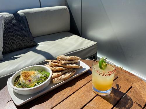 A plate of flatbreads next to a bowl of salad and a cocktail on a wooden table, with a cozy couch in the background.